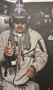 Man in grey sweats wearing a Happy New Year party hat, dark goggles, earmuff style hearing protection, holding a bottle of sparkling wine